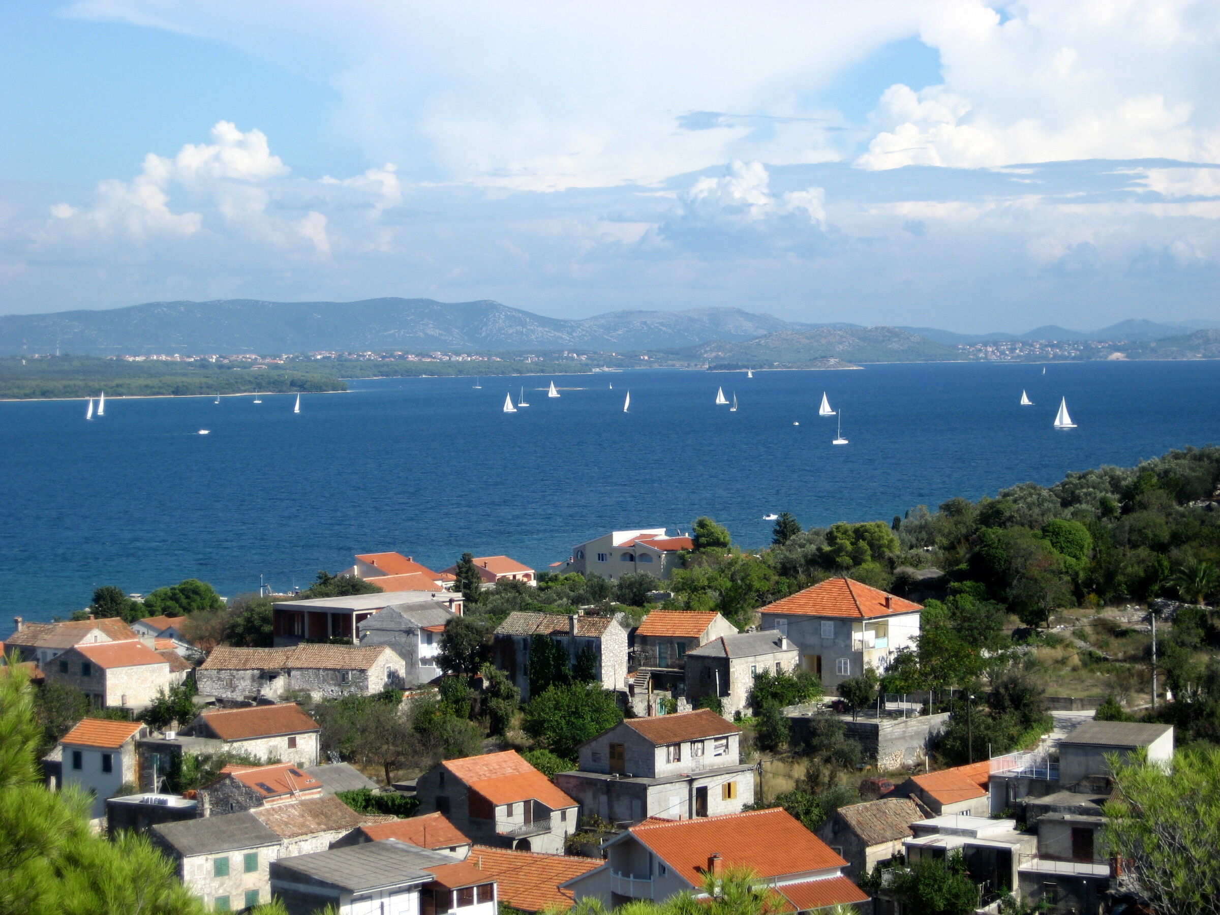 Red-roofed stone houses of Tkon on Pašman above a blue channel full of sailboats, with mainland mountains behind